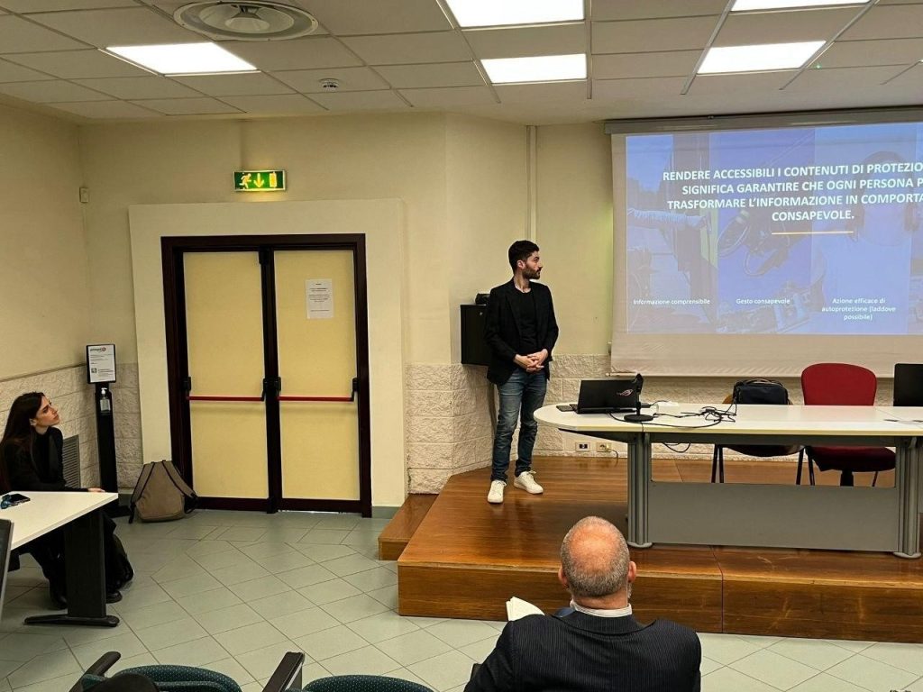 Evento Roma Tor Vergata il relatore sordo è sul palco e presenta i contenuti delle slide proiettate alle sue spalle; alla sua destra, un’interprete di Lingua dei Segni è seduta e traduce il suo intervento per il pubblico